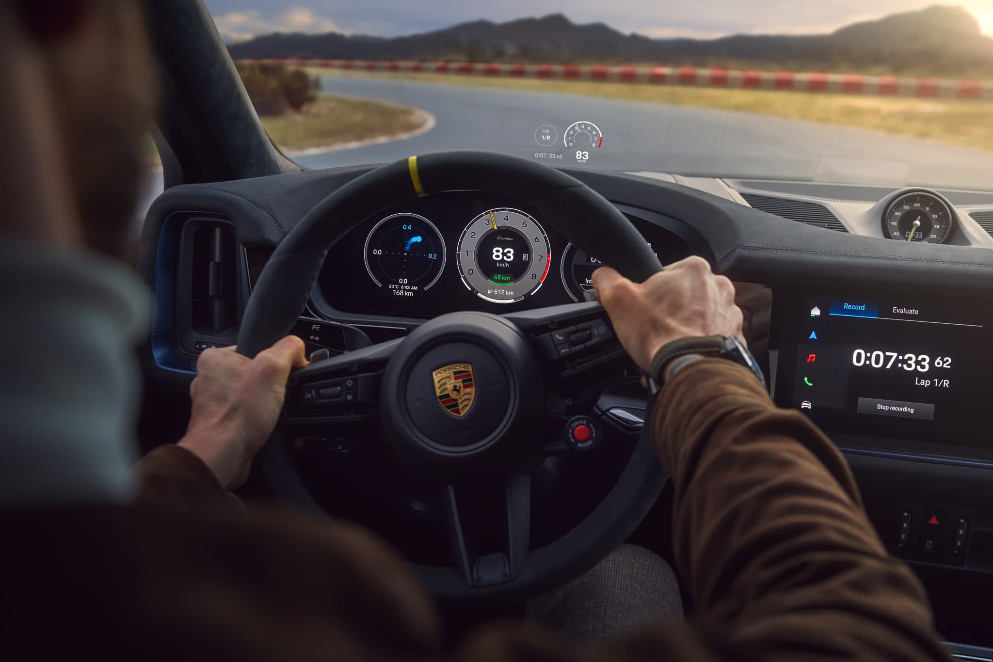 Driver steering a Cayenne Turbo E-Hybrid Coupe on a racetrack, dashboard displays speed and lap time, with mountains in the background.